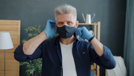 Portrait-of-serious-guy-putting-on-fabric-multiple-use-face-mask-looking-at-camera-in-house