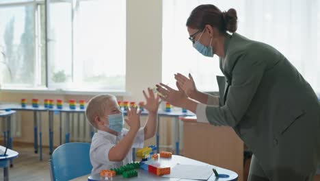 masked-little-cute-boy-with-down-syndrome-playing-pat-a-cake-with-her-teacher-sitting-at-desk,-leisure-in-school-after-lockdown