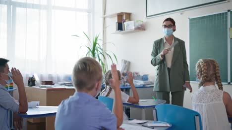 después-de-la-vida-de-cuarentena,-el-maestro-enmascarado-muestra-a-los-alumnos-tarjetas-con-cartas,-los-niños-levantan-la-mano-sabiendo-la-respuesta-correcta-sentado-en-los-escritorios-en-el-aula