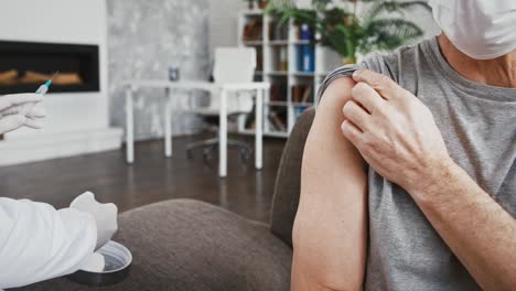 Female-doctor-in-disposable-gloves-injecting-vaccine-into-hand-of-senior-male-in-protective-mask-who-sitting-on-couch-at-home