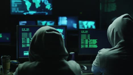 Two-hackers-in-hoods-work-on-a-computers-with-maps-and-data-on-display-screens-in-a-dark-office-room.