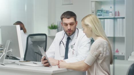 In-Doctor's-Office-Professional-Doctor-Consults-Beautiful-Young-Woman-with-a-Help-of-a-Tablet.-They-Look-and-the-Screen-and-Smile.