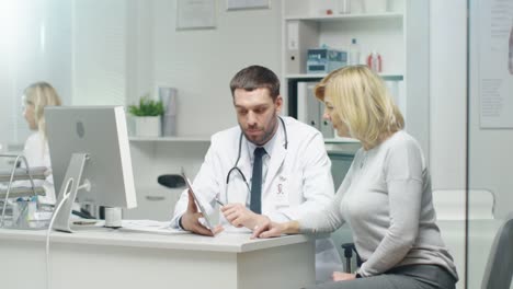 Professional-Doctor-Consults-Mid-Adult-Woman.-He-Points-out-Something-on-the-Screen-to-Her.-Nurse-Puts-Documents-on-Doctor's-Table.