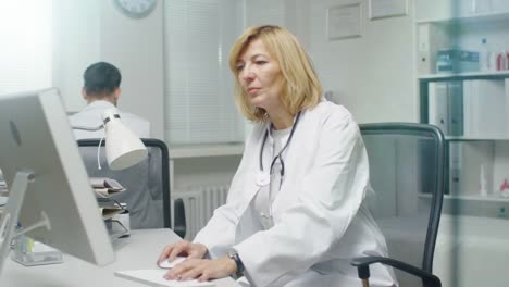 In-the-Doctor's-Office.-Mid-Adult-Female-Doctor-Works-at-Her-Desktop-Computer.-Her-Assistent-Stands-Up-and-Leaves-the-Office.