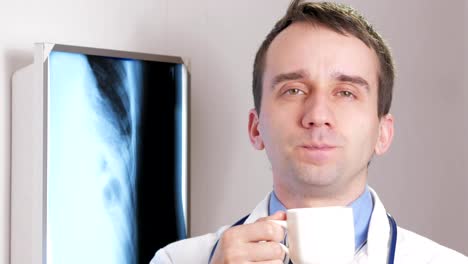 A-young-doctor-looking-at-the-camera-is-drinking-a-hot-drink-from-a-white-mug.-On-the-neck-hangs-a-stethoscope.-Close-up