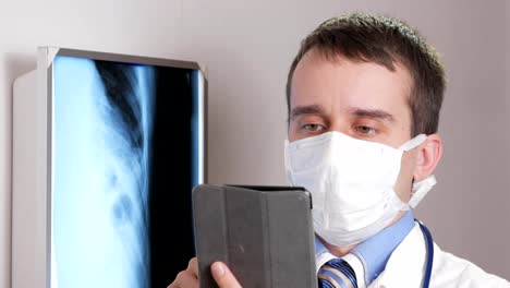 Ein-junger-Arzt-in-eine-Gesichtsmaske-verwendet-einen-Tabletcomputer-in-der-Klinik.-Ein-Mann-hält-die-Ergebnisse-der-Röntgenaufnahme-eines-Patienten