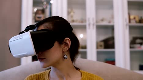 Vista-cercana-de-joven-mujer-en-gafas-de-realidad-virtual-sentado-en-el-sofá-en-casa.-Mujer-mirando-a-su-alrededor-en-VR-casco-con-auriculares