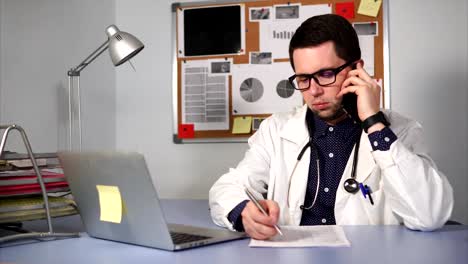 Male-medic-in-the-clinic-sitting-at-the-desk-and-talking-by-smartphone.