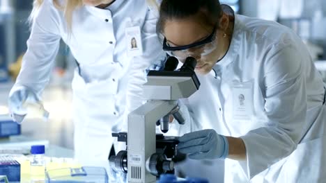 Medical-Research-Scientist-Drops-Sample-on-Slide-and-Her-Colleague-Examines-it-Under-Microscope.-They-Work-in-a-Modern-Laboratory.