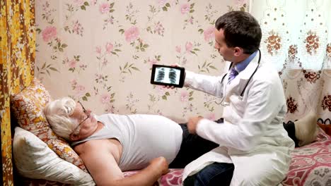 A-male-doctor-visits-a-patient-at-home.-It-shows-the-results-of-the-X-ray-on-a-tablet-computer.-The-man-is-lying-on-the-couch