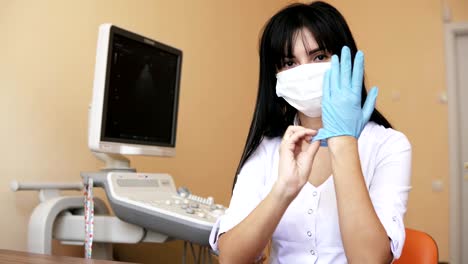 Attractive-smiling-female-doctor-in-surgical-mask-dressing-medical-gloves-while-sitting-at-the-table-in-her-office.-Shot-in-4k