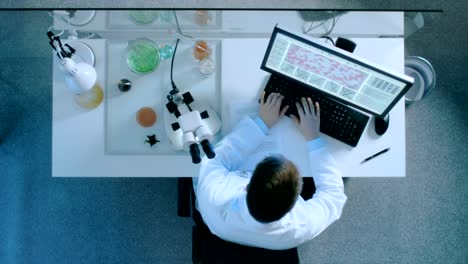 Top-Down-View-of-the-Research-Scientists-Looking-Under-Microscope-and-Entering-New-Data-in-the-Personal-Computer.