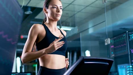 In-Scientific-Sports-Laboratory-Beautiful-Woman-Athlete-Walks-on-a-Treadmill-with-Electrodes-Attached-to-Her-Body,-Monitors-Show-EKG-Data-on-Display.-Slow-Motion.-Low-Angle.