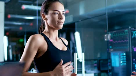 In-wissenschaftlichen-Sport-Labor,-die-schöne-Sportlerin-auf-einem-Laufband-mit-Elektroden-an-ihrem-Körper-geht,-anzeigen-Monitore-EKG-Daten-auf-dem-Display.-Slow-Motion.-Niedrigen-Winkel.