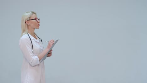 Blonde-Doctor-writing-on-a-clipboard-while-smiling-against-a-grey-background