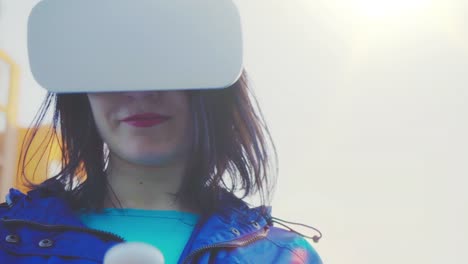 Mujer-joven-con-gafas-VR,-auriculares,-jugar-juegos-virtuales-y-ver-vídeos-al-aire-libre-contra-el-fondo-de-un-edificio-de-cristal