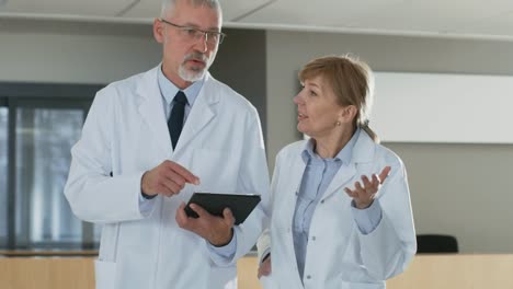 In-the-Hospital-Doctors-Walking-through-Lobby,-have-Discussion-while-Using-Tablet-Computer.-In-the-Background-Patients-and-Medical-Personnel.-New-Modern-Fully-Functional-Medical-Facility.