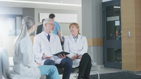 In-the-Lobby-of-the-Hospital-Three-Professional-Doctor-Have-Discussion-while-Sitting.-They-Use-Tablet-Computer.-Busy-Modern-Hospital-with-Best-Specialists-and-Medicare-in-Country.