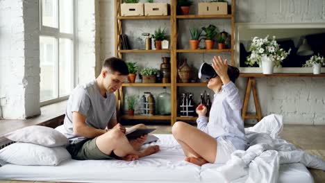 Married-couple-is-having-fun-with-augmented-reality-glasses,-young-woman-is-wearing-then-and-smiling-moving-hands-and-her-husband-is-using-tablet.-Technology-and-people-concept.