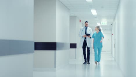 Surgeon-and-Female-Doctor-Walk-Through-Hospital-Hallway,-They-Consult-Digital-Tablet-Computer-while-Talking-about-Patient's-Health.-Modern-Bright-Hospital-with-Professional-Staff.