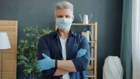 Retrato-de-hombre-adulto-con-máscara-médica-preventiva-y-guantes-de-pie-en-el-apartamento-con-los-brazos-cruzados