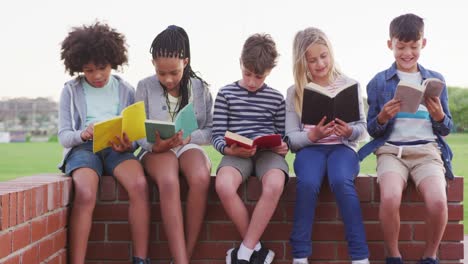 Grupo-de-niños-leyendo-libros-mientras-están-sentados-en-una-pared-de-ladrillos