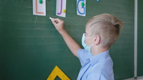 Schuljunge-in-medizinischer-Maske-in-der-Nähe-von-Tafel-schreibt-in-Kreide-die-Buchstaben-in-Klassenzimmer,-Schule-nach-Quarantäne-und-Sperrung