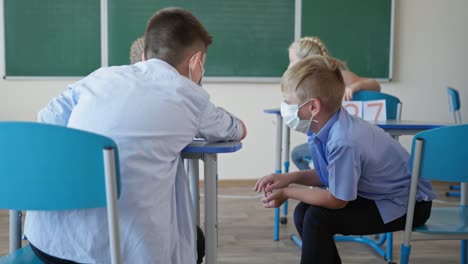 compañeros-de-clase-enmascarados-en-los-escritorios-traseros-están-hablando-entre-sí-durante-la-clase-o-romper-en-el-fondo-de-la-pizarra-en-la-escuela