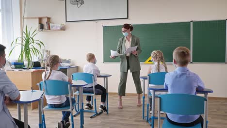 Schule-nach-Quarantäne,-Lehrer-in-medizinischer-Maske-verteilt-Flugblätter-mit-Auftrag-an-Schüler-sitzen-am-Schreibtisch-während-einer-Unterrichtsstunde-im-Klassenzimmer