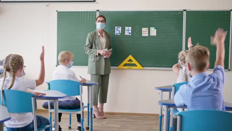 study-during-pandemic,-teacher-in-medical-mask-and-glasses-at-lesson-near-whiteboard-asks-pupils-questions,-schoolgirl-is-ready-to-answer-and-raises-hand