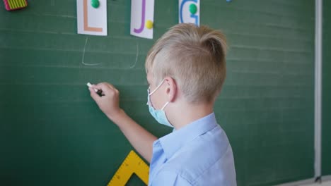 Schüler-in-medizinischer-Maske-an-der-Tafel-schreibt-Briefe-mit-Kreide-im-Klassenzimmer-in-der-Schule-nach-Quarantäne-und-Sperrung