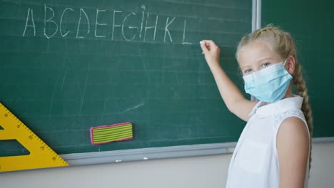 Portait-von-Schülerin-in-medizinischer-Maske-schreibt-in-Kreide-das-englische-Alphabet-im-Klassenzimmer,-Schüler-Blick-auf-die-Kamera-in-der-Nähe-von-Tafel-in-der-Schule