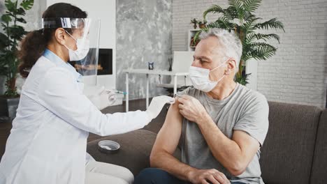 Mujer-médico-con-máscara-protectora-y-protector-facial-está-inyectando-vacuna-en-el-hombro-del-hombre-de-edad-que-se-sienta-en-el-sofá-en-casa