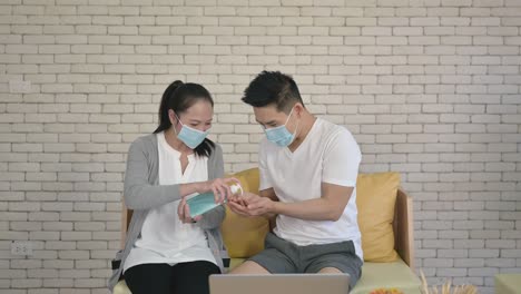 Asian-adult-couple-sitting-on-couch-working-at-home-wearing-hygiene-face-mask-and-wash-hands-by-alcohol-gel-or-sanitizer-to-protective-pandemic-of-coronavirus-or-covid-19.