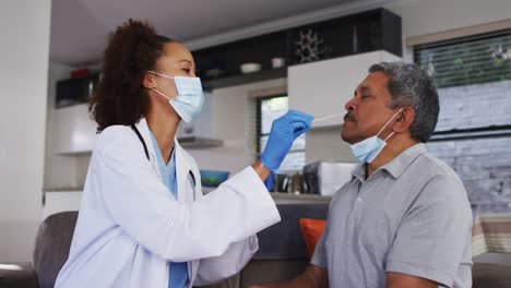 Mixed-race-female-doctor-wearing-mask-doing-swab-test-on-senior-man-at-home