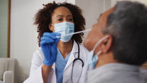 Mixed-race-female-doctor-wearing-mask-doing-swab-test-on-senior-man-at-home