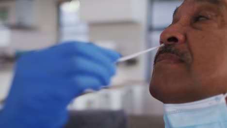 Mixed-race-female-doctor-wearing-mask-doing-swab-test-on-senior-man-at-home