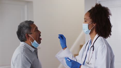 Mixed-race-female-doctor-wearing-mask-doing-swab-test-on-senior-man-at-home