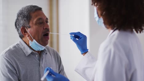 Mixed-race-female-doctor-wearing-mask-doing-swab-test-on-senior-man-at-home