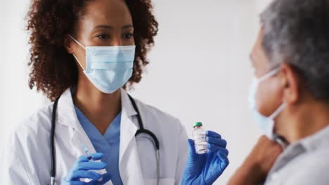 Mixed-race-female-doctor-wearing-mask-giving-vaccination-to-senior-man-at-home