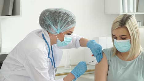 Medical-nurse-in-gloves-and-protective-mask-vaccinates-female-patient-in-clinic.-Doctor-injects-vaccine-against-Covid-19-into-hand-from-syringe.-Coronavirus