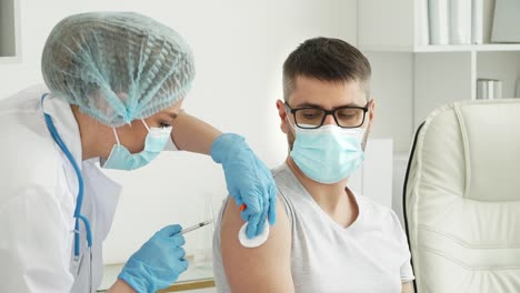 Medical-nurse-in-safety-gloves-and-protective-mask-vaccinates-male-patient-in-clinic.-Doctor-injects-medicine-into-hand-from-syringe-and-applies-medical-patch.-Covid-19,-coronavirus-vaccine