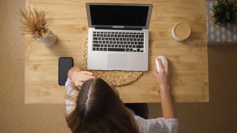 The-girl-works-remotely-via-the-Internet.-He-sits-at-his-laptop-and-presses-the-mouse.-Top-view.-Wooden-table.-Beige-colors.-The-concept-of-e-learning-from-home.-Selective-Focus.