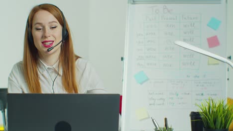 Woman-in-headphones-talking-to-students-via-video-conference-at-empty-class.-Smiling-female-school-teacher-having-online-class-indoors.-Education-concept.-Quarantine-concept.-Study-by-internet.