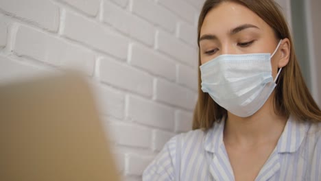 Self-isolation.-A-girl-in-a-medical-mask-and-striped-pajamas-close-up.-Works-remotely.-Prints-text-on-a-laptop.