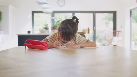 Young-Asian-Girl-Home-Schooling-Working-At-Table-In-Kitchen-Writing-In-Book-During-Lockdown