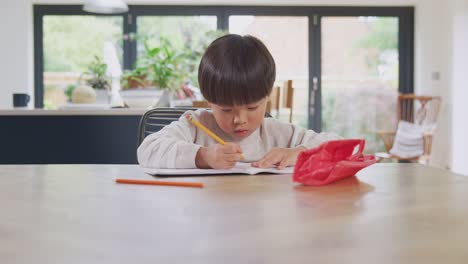 Young-Asian-Boy-Home-Schooling-Working-At-Table-In-Kitchen-Writing-In-Book-During-Lockdown