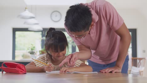 Asiatische-Vater-hilft-Zu-Hause-Schulende-Tochter-arbeiten-am-Tisch-in-Küche-Schreiben-in-Buch