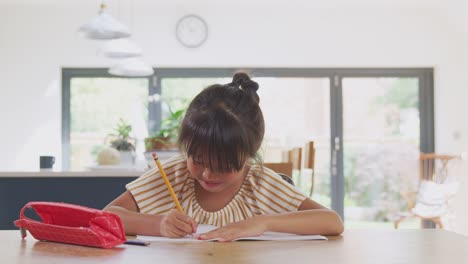 Young-Asian-Girl-Home-Schooling-Working-At-Table-In-Kitchen-Writing-In-Book-During-Lockdown