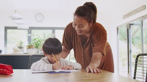 Asian-Mother-Helping-Home-Schooling-Son-Working-At-Table-In-Kitchen-Writing-In-Book